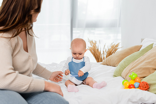 Young body positive mother sitting near baby with plate and spoon on bed