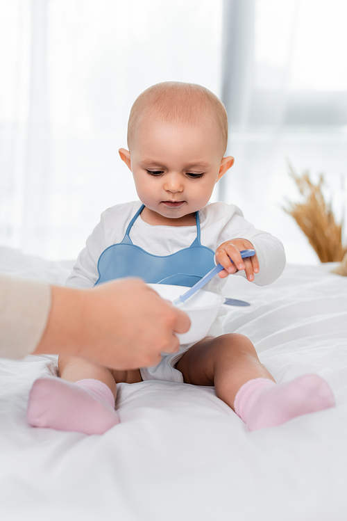 Woman holding plate with baby food near daughter with spoon on bed