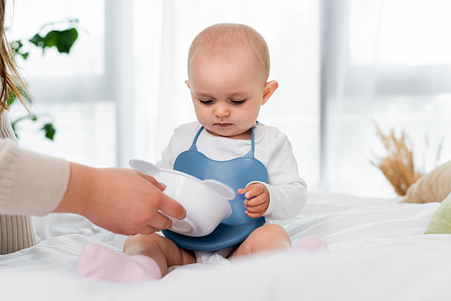 Woman holding plate near baby daughter on bed at home