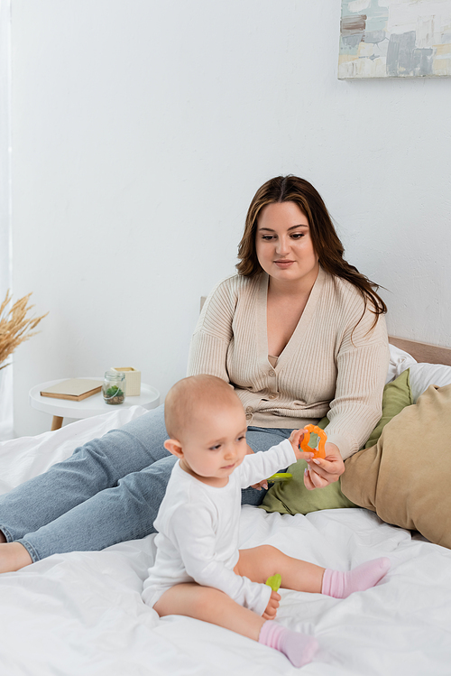 Young plus size woman looking at baby daughter with toys on bed