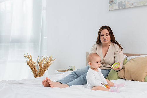 Plus size mother holding toy near baby girl on bed at home