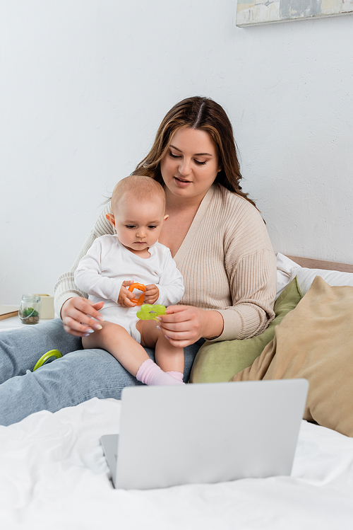 Young body positive woman holding toy near baby and laptop on bed