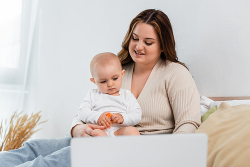 Smiling plus size mother looking at child with toy near blurred laptop on bed