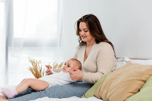 Positive plus size woman touching hands of baby daughter on bed