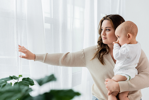 Young body positive mother holding child near curtains at home