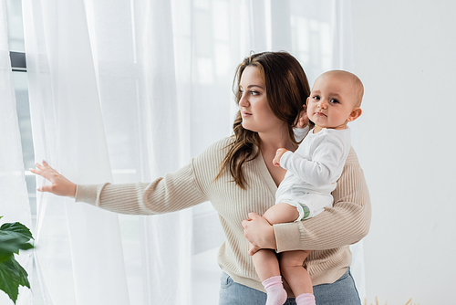 Pretty woman with overweight holding baby near curtains at home