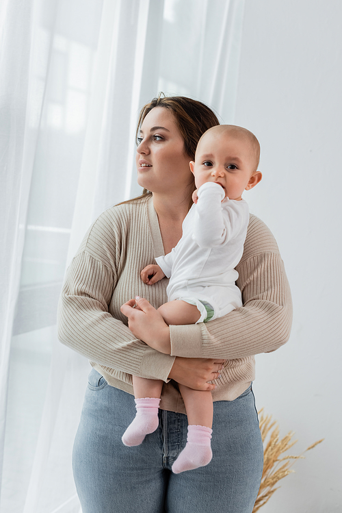 Brunette body positive woman holding baby girl near curtains at home