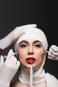 plastic surgeons in latex gloves holding surgical knife and syringes near woman in elastic bandage on head isolated on dark grey