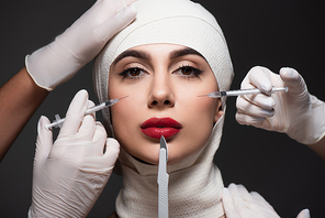 plastic surgeons holding surgical knife and syringes near patient in elastic bandage on head isolated on dark grey