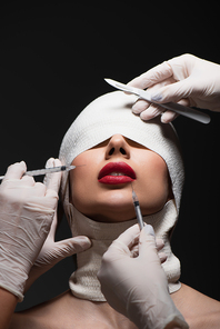 young patient in elastic bandage near plastic surgeons with surgical knife and syringes isolated on grey