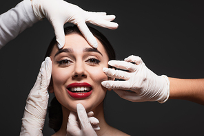 plastic surgeons in latex gloves touching face of smiling young woman isolated on black