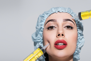 huge syringes near face of young woman with red lips and surgical cap isolated on grey