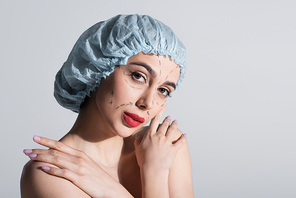 young woman in blue surgical cap with marked lines on face embracing bare shoulders isolated on grey