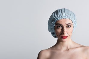 serious young woman in surgical cap with marked lines on face isolated on grey