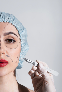 cropped view of plastic surgeon in latex glove holding scalpel near woman with marked lines on face isolated on grey