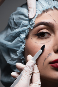 close up of plastic surgeon in latex gloves holding scalpel near woman with marked lines on face isolated on dark grey