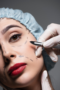 close up of plastic surgeon in latex gloves holding scalpel near woman with and marked lines on face isolated on grey