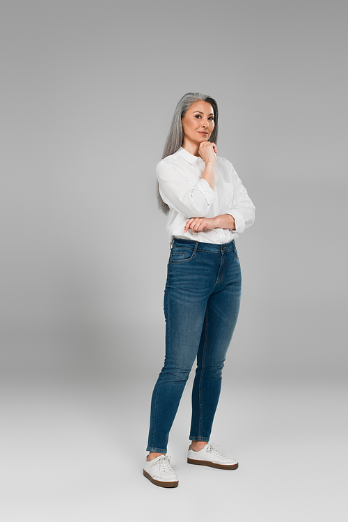 full length view of mature woman in white shirt and jeans standing with hand near chin on grey background