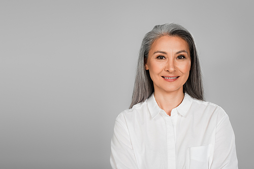 joyful middle aged woman in white shirt smiling at camera isolated on grey