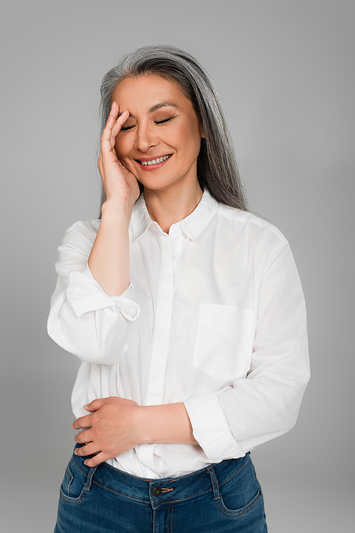 mature woman in white shirt touching face while laughing with closed eyes isolated on grey