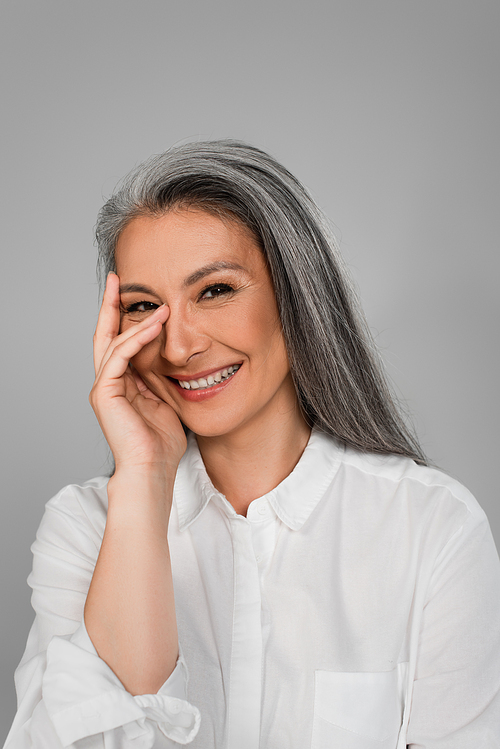 cheerful middle aged woman holding hand near face while smiling at camera isolated on grey