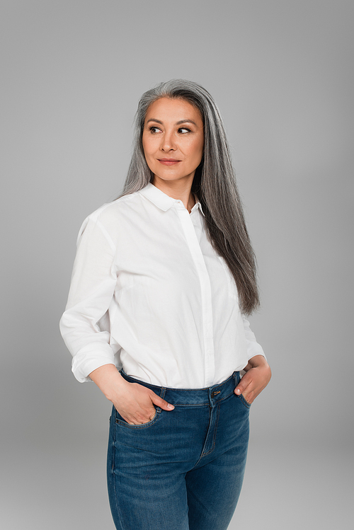 middle aged woman standing with hands in pockets of jeans isolated on grey