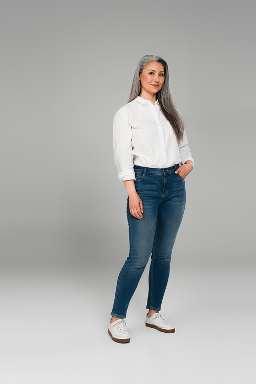 full length view of smiling middle aged woman in blue jeans and white shirt on grey