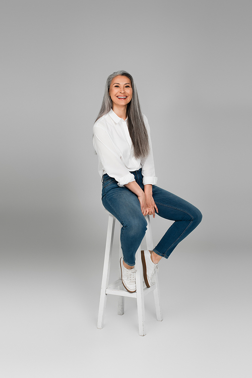 cheerful middle aged woman in blue jeans and white shirt sitting on high stool on grey background
