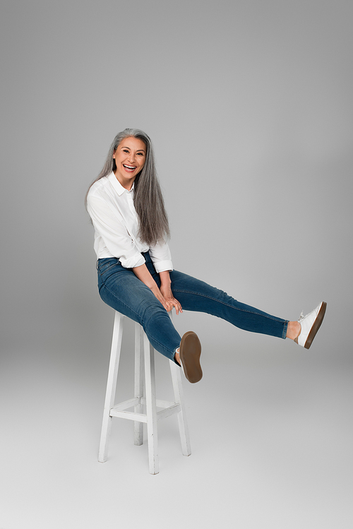 mature woman in blue jeans and white shirt laughing on high stool on grey background