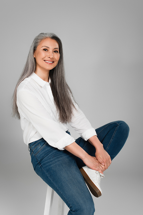 happy middle aged woman wearing blue jeans and white shirt, sitting on stool isolated on grey