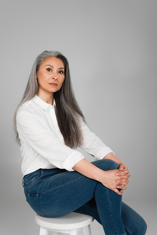 grey haired woman in white shirt and jeans sitting on stool isolated on grey