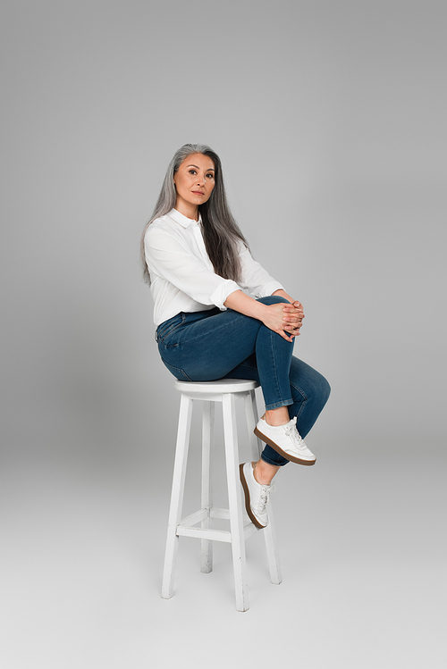 grey haired woman in white shirt and blue jeans  on high stool on grey background