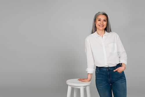smiling middle aged woman in white shirt standing near high stool with hand in pocket of jeans isolated on grey