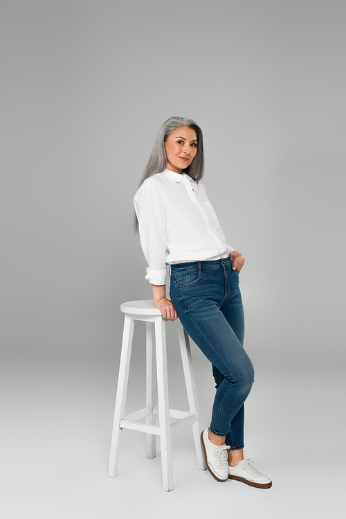 stylish middle aged woman in white shirt standing with hand in pocket of jeans near high stool on grey background