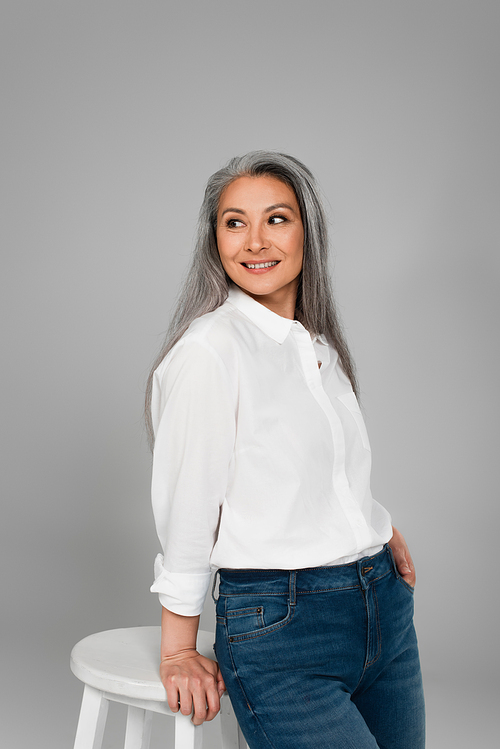 positive mature woman in white shirt standing with hand in pocket of jeans isolated on grey