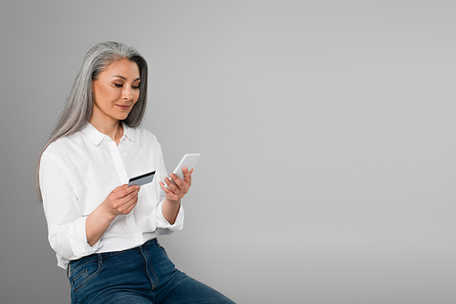 stylish middle aged woman sitting with credit card and mobile phone isolated on grey