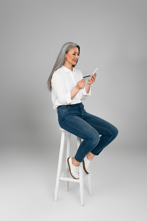 grey haired woman in jeans and shirt sitting on high stool with cellphone and credit card on grey background
