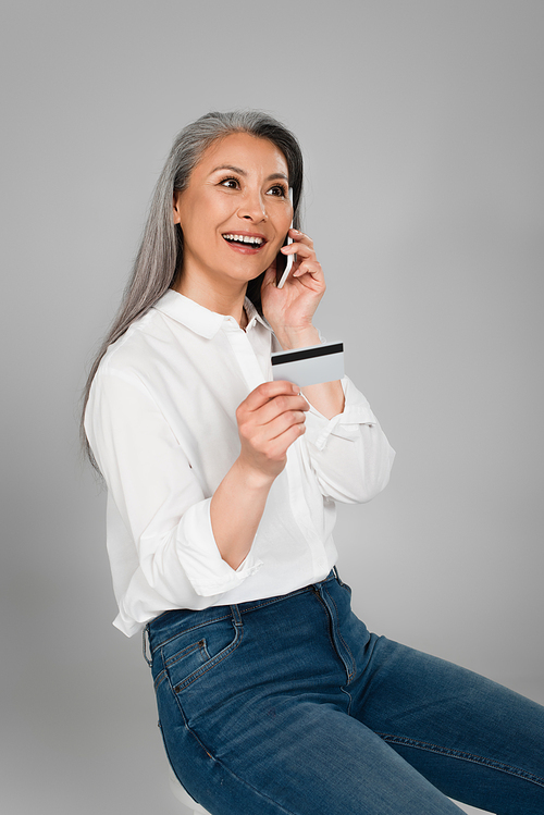 happy mature woman with credit card talking on mobile phone isolated on grey
