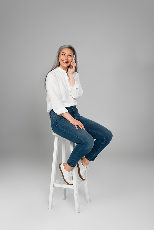 cheerful middle aged woman in jeans sitting on high stool and talking on smartphone on grey background
