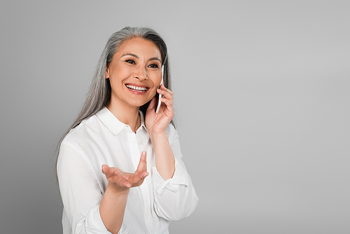 mature woman in white shirt smiling and gesturing while talking on mobile phone isolated on grey