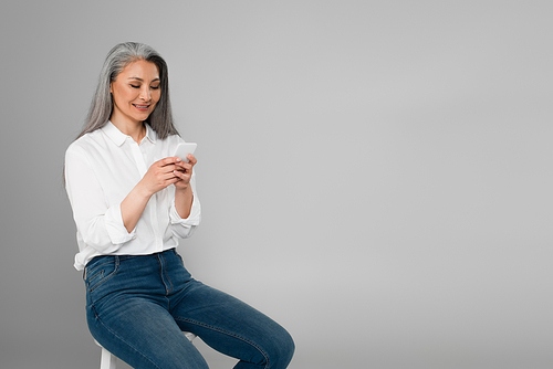 happy and stylish middle aged woman messaging on smartphone while sitting isolated on grey