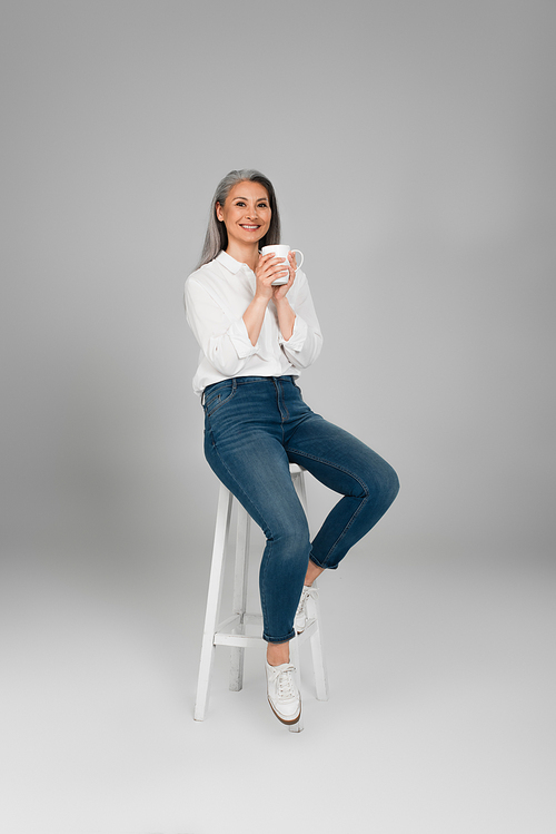 happy mature woman in shirt and jeans sitting on high stool with tea on grey background