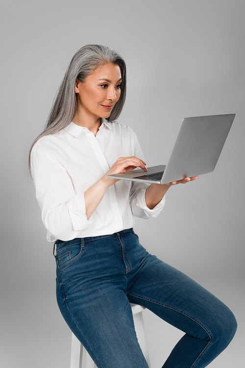 middle aged woman with grey hair sitting on stool and typing on laptop isolated on grey
