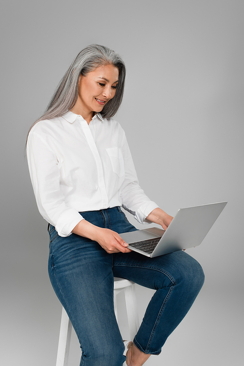 happy mature woman in white shirt typing on laptop while sitting on stool isolated on grey