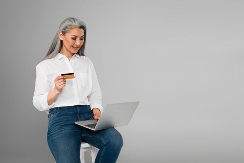 smiling middle aged woman with credit card sitting on stool and using laptop isolated on grey