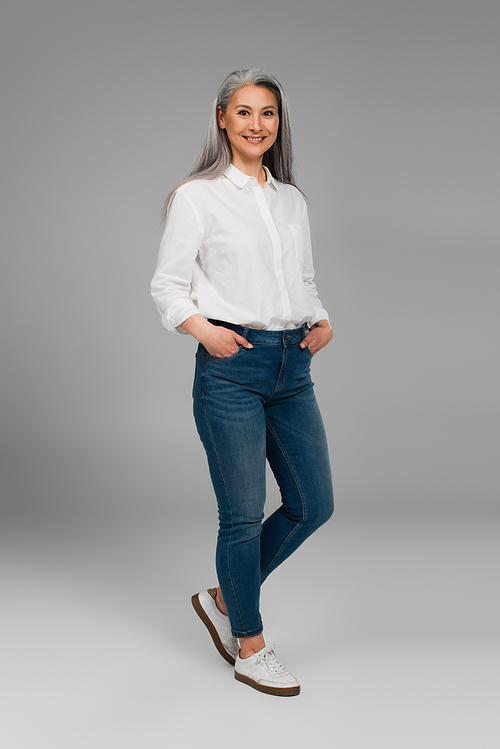 full length view of middle aged woman with hands in pockets of blue jeans smiling on grey