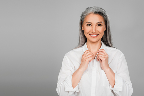 pleased mature woman adjusting collar of white shirt while  isolated on grey