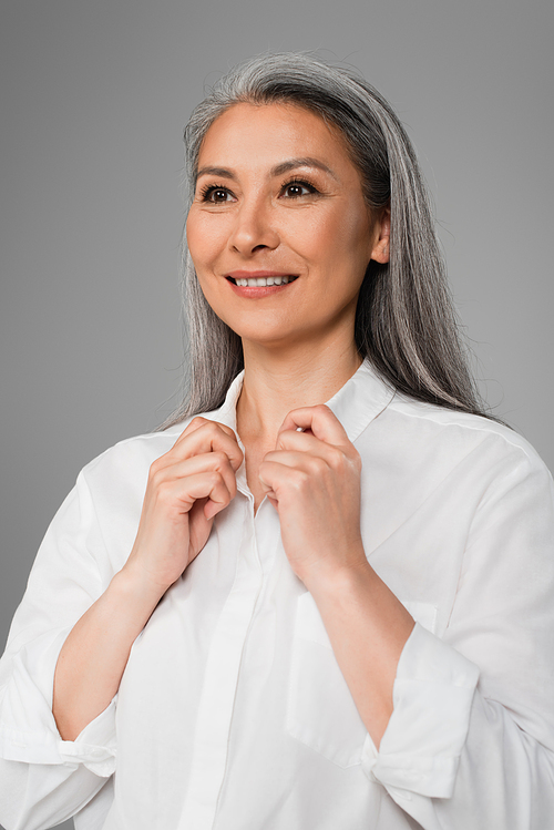 mature woman smiling while touching collar of white shirt isolated on grey