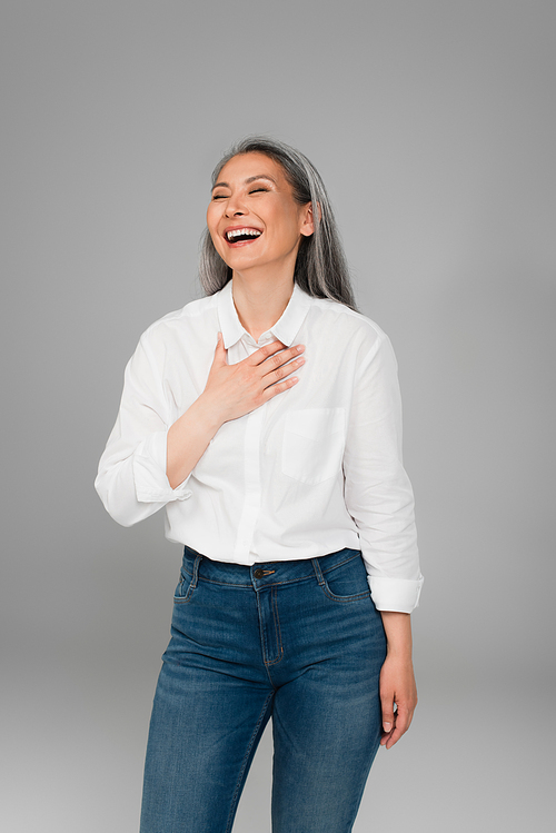 excited mature woman touching chest while laughing with closed eyes isolated on grey