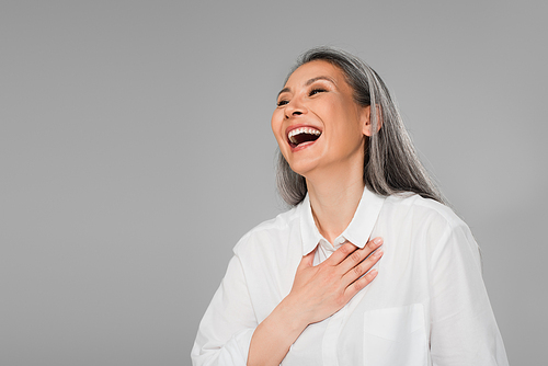 overjoyed woman with grey hair touching chest while laughing isolated on grey